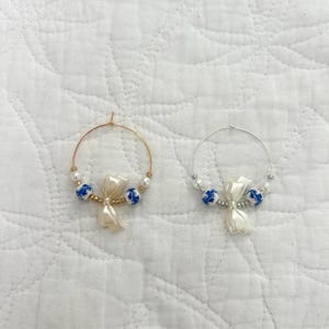 May include: Two hoop earrings. One is gold-toned, the other silver-toned. Each earring features a white bow-shaped charm, blue and white porcelain beads, and small white beads. The earrings are displayed on a white, textured surface.
