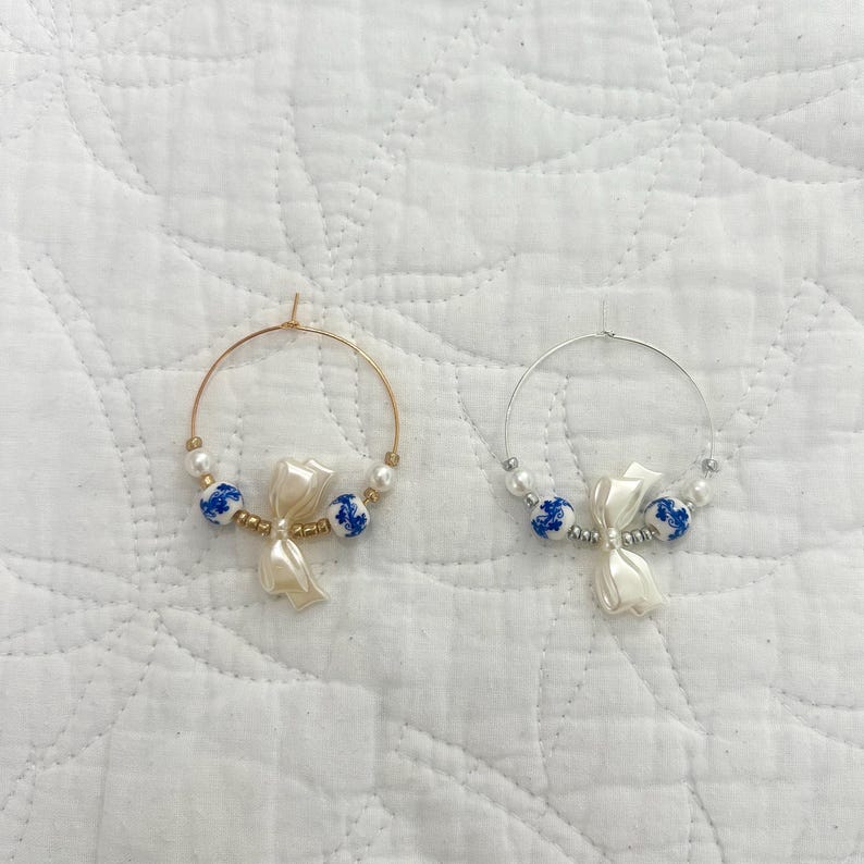 May include: Two hoop earrings. One is gold-toned, the other silver-toned. Each earring features a white bow-shaped charm, blue and white porcelain beads, and small white beads. The earrings are displayed on a white, textured surface.