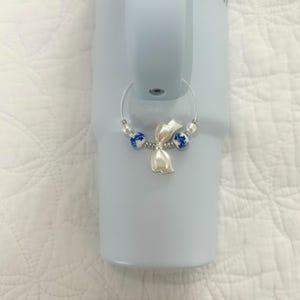 May include: A silver wine glass charm with a white flower and blue and white beads. The charm is attached to a light blue tumbler. The beads are round and the flower is a stylized tulip.