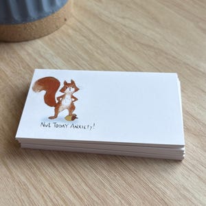 Puede incluir: Una pila de tarjetas blancas con una ilustración en acuarela de una ardilla sosteniendo una bellota. La ardilla es marrón con una cola tupida. El texto "Nut Today Anxiety!" está impreso debajo.
