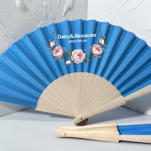 May include: A blue folding fan with a floral design and the text "Daisy & Alexander 2025/05/25". The fan is made of paper and wood, with a light blue pleated design and a light wood handle.