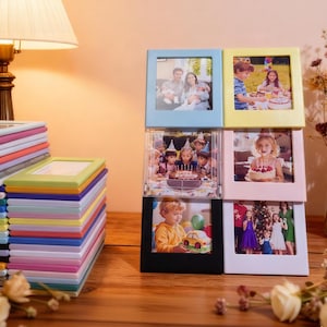 May include: A stack of colorful photo frames in various colors, including pink, green, and blue, next to a display of framed photos. The frames are arranged on a wooden surface, with a lamp and dried flowers in the background.