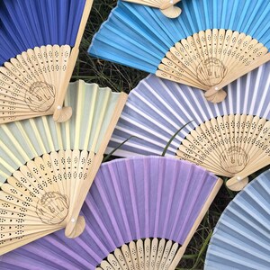 Puede incluir: Un conjunto de abanicos de mano abiertos en tonos azules, morados y amarillos. Los abanicos tienen mangos de madera y están decorados con un delicado diseño. Están colocados sobre una superficie cubierta de hierba.
