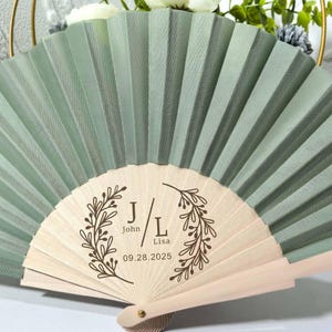 May include: A sage green folding fan with a wooden handle. The fan features the initials "J/L", the names "John" and "Lisa", and the date "09.28.2025" within a decorative wreath design. The fan is open, displaying its pleated design.