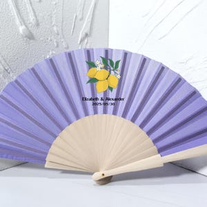 May include: A lilac hand fan with a lemon and leaf design. The fan has a wooden handle and features the text "Elizabeth & Alexander 2025/05/30". The fan is open, displaying its pleated design.