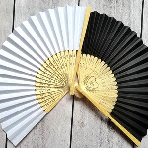 May include: Two paper folding fans, one white and one black, with a wooden handle and a decorative design featuring a heart and the initials A&B. The fans are open and facing each other.