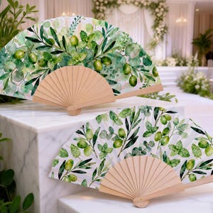 May include: Two open hand fans with a watercolor design of green olives and leaves. The fans have a natural wood handle and are displayed on a white marble surface. The background shows a wedding ceremony setting.