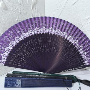 May include: A decorative purple hand fan with a white lace design. The fan is open, displaying a semi-circular shape. The fan is made of wood and fabric. The text "Victoria & William 2025-08-05" is printed on the fan. Several closed fans are below.