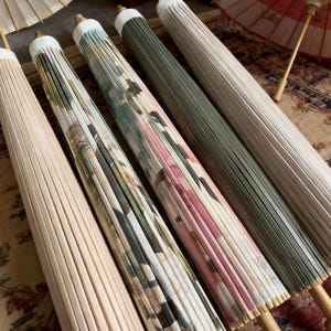 Puede incluir: Cinco sombrillas cerradas dispuestas una al lado de la otra. La primera es de color crema liso, la segunda presenta un estampado botánico verde y blanco, la tercera muestra un patrón floral rosa y verde, la cuarta es verde oliva y la quinta es blanca.