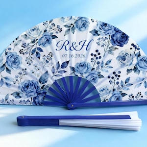 May include: A white and blue floral hand fan with the initials "R&H" and the date "07.16.2026" printed on it. The fan features a blue floral design and a blue handle. A second, folded fan is below.