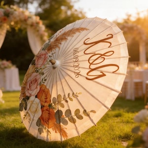 May include: A cream-colored paper parasol featuring a floral print of orange, peach, and white roses with green eucalyptus leaves and pampas grass. The parasol is personalized with the brown script letters K&G and the date 06.09.2026.