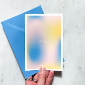 Op de afbeelding: Een wenskaart met een zachte, vage achtergrond in blauw, geel en roze tinten, met de woorden "welcome home" in een sierlijk lettertype. De kaart ligt naast een blauwe envelop.