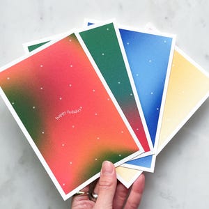 Könnte beinhalten: Eine Hand hält einen Fächer aus farbenfrohen Grußkarten. Jede Karte hat einen Farbverlaufshintergrund in Rot, Grün, Blau und Gelb mit weißen Sternen und dem Text "happy holiday".