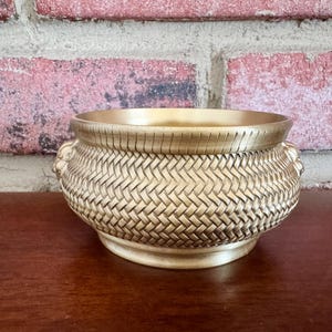 May include: A gold-colored decorative bowl with a woven basket design. The bowl has a wide opening and a rounded base. The exterior features a textured, basket-weave pattern. The bowl is sitting on a dark brown surface, with a brick wall in the background.