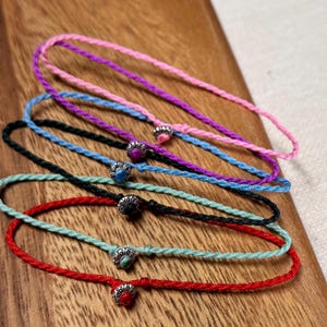 May include: A collection of six braided cord bracelets in various colors: pink, purple, blue, black, green, and red. Each bracelet features a small, decorative silver-toned bead. The bracelets are displayed on a wooden surface.