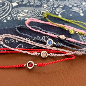 May include: A collection of colorful friendship bracelets. Each bracelet features a central gemstone in a silver setting. The bracelets are in shades of red, pink, blue, and green, with a braided cord design.