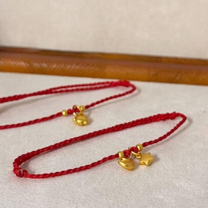 May include: Two red braided bracelets with gold-colored charms. One bracelet has a charm shaped like a small cat, and the other has a cat and a star charm. The bracelets are displayed on a light-colored surface.