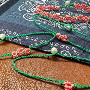 May include: A collection of handmade bracelets featuring green cord, small pink flower-shaped knots, and white spherical beads. The bracelets are constructed with thin, knotted green string and include delicate floral accents made from pink thread.