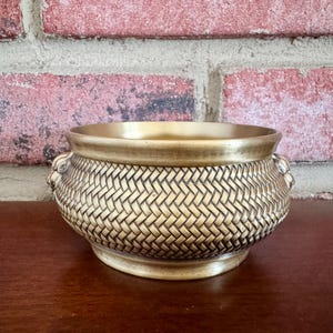 May include: A gold-colored decorative bowl with a woven basket pattern. The bowl has a wide rim and two small handles on either side. The background features a brick wall.