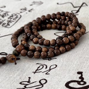 May include: A strand of dark brown wooden beads, likely a prayer mala or meditation bracelet, is coiled on a light-colored fabric. The beads are round and appear to be made of a natural wood material. The background fabric has a subtle pattern.