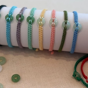 May include: A set of ten bracelets with green jade beads and different colored cords. The bracelets are displayed on a white surface.