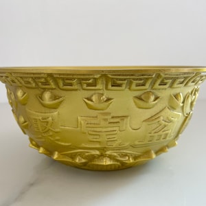 May include: A gold-colored decorative bowl with intricate embossed designs. The bowl features symbols and characters, possibly of Asian origin, and is set against a white background. The bowl's rim has a decorative border.