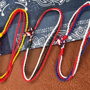 May include: Three handmade cord bracelets featuring knotted closures. The first is yellow and red, the second is black, white, and red, and the third is blue, white, and red. Each bracelet is constructed from thin, twisted fibre strands.
