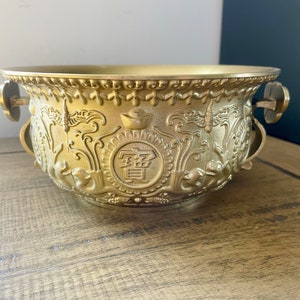 May include: A gold-colored, ornate bowl with handles, featuring detailed relief carvings of dragons, symbols, and Chinese characters. The bowl has a wide rim and a rounded body, suggesting a decorative or ceremonial purpose.