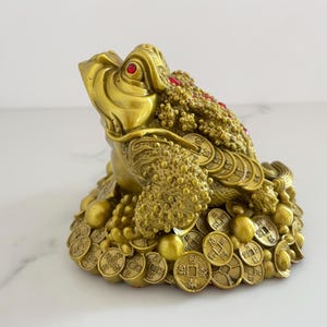 May include: A golden, detailed frog statue with red gem eyes and coins. The frog is covered in textured bumps and sits atop a base of coins and golden spheres. This decorative item is a symbol of wealth and prosperity.