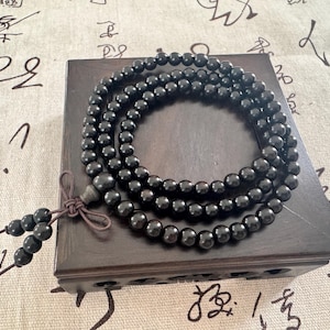 May include: A dark wooden prayer bead necklace with a tassel, resting on a dark wooden box. The beads are round and polished, and the necklace is arranged in a circular pattern. The background is a patterned fabric.