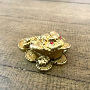 May include: A golden frog figurine with red gem eyes, resting on a bed of gold coins. The frog is detailed with textured skin and sits on six coins. The figurine is displayed on a wooden surface.