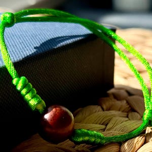 May include: A green string bracelet with a dark brown wooden bead. The bracelet is tied with a knot on one end and has a loose end on the other.