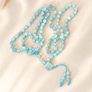 May include: A long necklace featuring translucent light blue spherical beads strung on a matching light blue cord. The necklace includes a central square-shaped light blue bead and terminates in two short dangling strands with small beads.