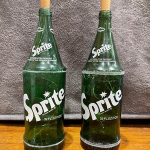 May include: Two green glass Sprite soda bottles with white labels and a star logo. The bottles are 32 fluid ounces and have a return for deposit label. The bottles have cork stoppers.