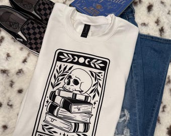 Puede incluir: Sudadera blanca con un diseño de carta de tarot en blanco y negro. La carta representa una calavera sobre una pila de libros con el texto "THE READER" debajo.