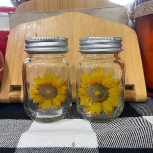 May include: Two clear glass jars with silver lids and sunflower decals. The jars are sitting on a black and white checkered surface.