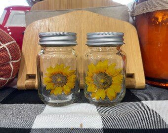 May include: Two clear glass jars with silver lids and sunflower decals. The jars are sitting on a black and white checkered surface.