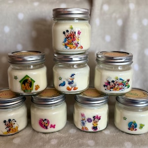 May include: Eight glass jars with lids, filled with white wax and decorated with Disney character stickers. The jars are arranged in two rows of four. The stickers feature Mickey Mouse, Minnie Mouse, Donald Duck, Goofy, Pluto, and other Disney characters.