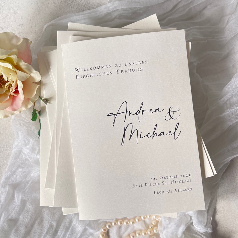 Pu&ograve; includere: Un programma di nozze bianco con testo nero. Il programma recita "Willkommen zu unserer kirchlichen Trauung" seguito dai nomi "Andrea & Michael". La data e la location del matrimonio sono elencate sotto i nomi: "14. Oktober 2023, Alte Kirche St. Nikolaus, Lech am Arlberg."
