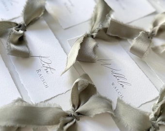Puede incluir: Tarjetas de lugar blancas con nombres escritos en caligrafía, atadas con lazos de cinta verde salvia. Las tarjetas de lugar tienen un borde áspero y desgarrado.