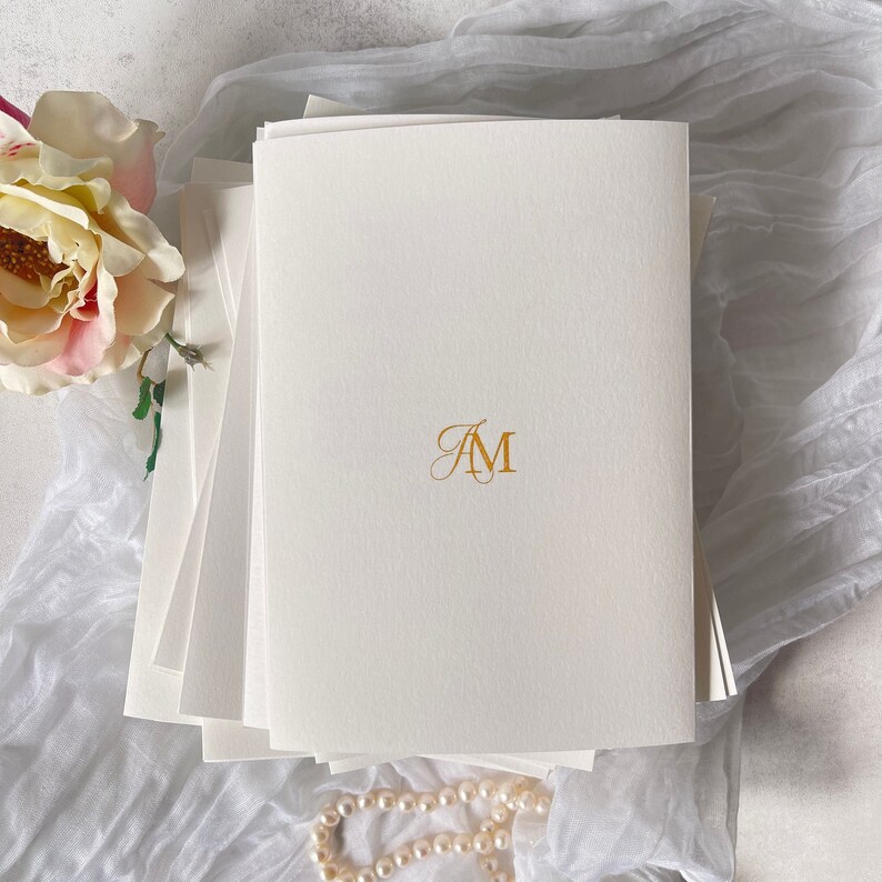 Pu&ograve; includere: Un mazzo di cartoncino bianco con un monogramma dorato "AM" stampato sul foglio superiore.