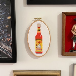 Puede incluir: Una bordado de punto de cruz de una botella de salsa picante roja con el nombre de la marca "Cholula" y la palabra "Original" en la etiqueta. La botella est&aacute; rodeada de un fondo blanco.