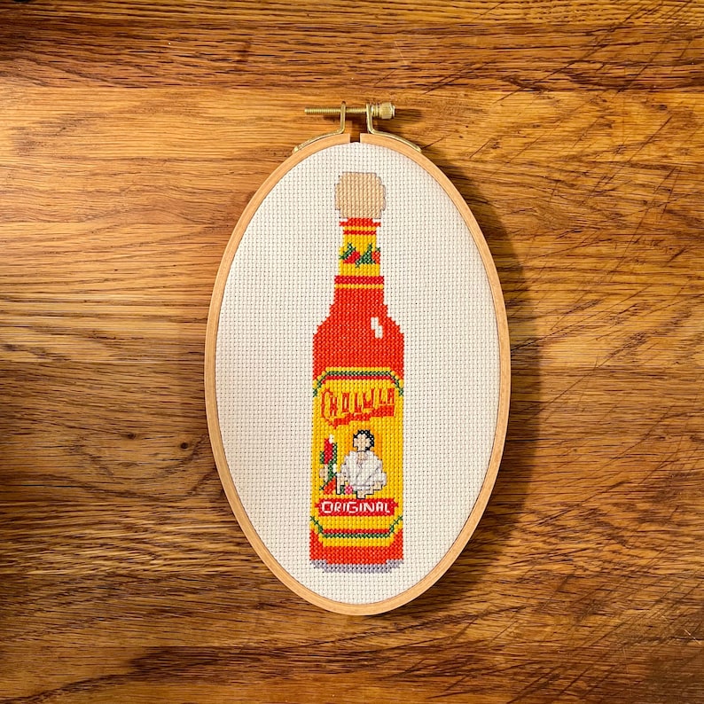Puede incluir: Una bordado de punto de cruz de una botella de salsa picante roja y amarilla con el nombre de la marca "Cholula" y la palabra "Original" en la etiqueta. La botella est&aacute; en un aro de bordado de madera.