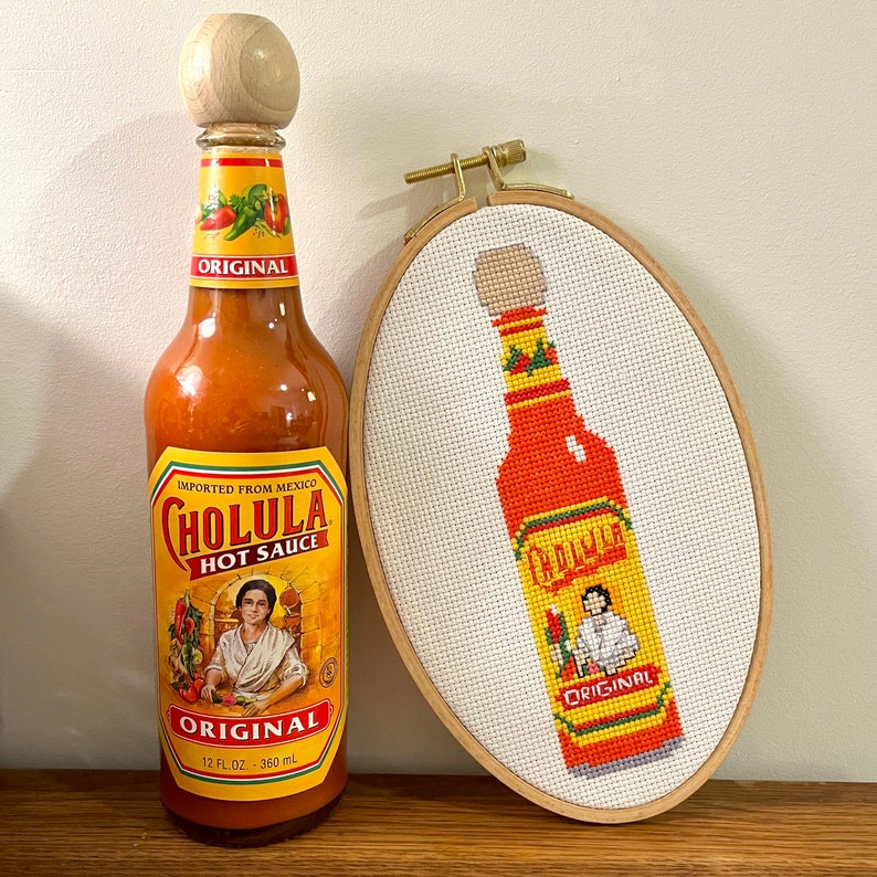 Puede incluir: Un bordado de punto de cruz de una botella de salsa picante roja y amarilla con el texto "Cholula Original" en la etiqueta. La botella est&aacute; enmarcada en un aro de madera. Una botella real de salsa picante Cholula Original est&aacute; en primer plano.