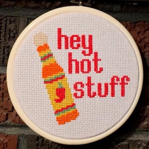 May include: A cross-stitch embroidery hoop featuring the phrase "hey hot stuff" in red letters. A colorful hot sauce bottle is stitched on the left side. The bottle is orange, yellow, and green with a red chili pepper.