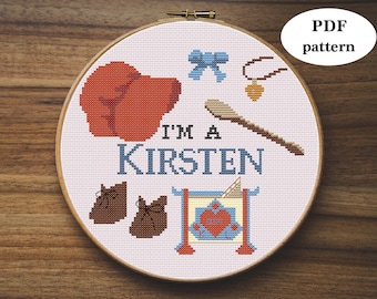 May include: A white embroidery hoop with a cross-stitch design featuring a red mitten, blue bow, brown shoes, a wooden spoon, a gold necklace, and the text "I'm a Kirsten".