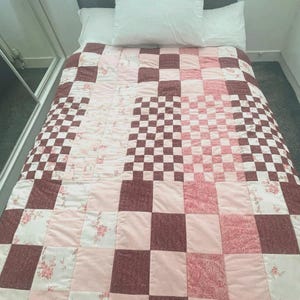 May include: A patchwork quilt in shades of pink and brown, arranged in a checkerboard pattern. The quilt features squares of solid colours, floral prints, and a checkerboard design. Two white pillows are on top.