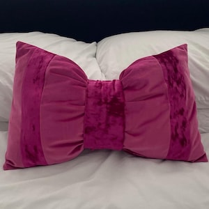 May include: A pink velvet pillow shaped like a bow. The pillow has a deep pink centre and lighter pink edges.