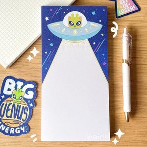 May include: A blue notepad with a white background and a cartoon alien in a spaceship. The spaceship is flying through a starry night sky with pink, blue, and yellow streaks. The notepad has a gold border and the word "avaira" is printed in the bottom right corner.