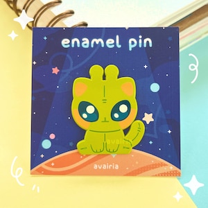May include: A green alien cat enamel pin with large eyes and a pink nose. The pin is set against a dark blue background with stars and a planet. The text "enamel pin" is at the top of the card and the text "avairia" is at the bottom.
